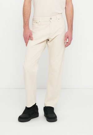 Pantalon en coton beige clair avec une coupe régulière, cinq poches et une fermeture par bouton. Associé à des baskets noires. Texture lisse.