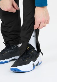 Svarta sportbyxor med dragkedjor vid bensluten lyfts för att avslöja en svart och vit sneaker med blå accenter samt vita ankelstrumpor.