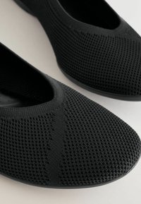 Zapatos negros sin cordones con parte superior de malla texturizada, presentan una abertura elástica y una suela plana y flexible.