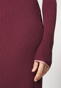 Main reposant à côté de la manche d'une robe en maille côtelée bordeaux avec un trou pour le pouce et une bordure contrastante rose clair.