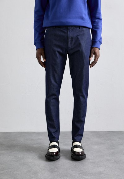 Dark blue denim trousers with a slim fit, featuring belt loops and side pockets. Paired with black and white footwear.