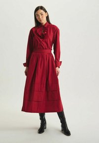 Robe en coton rouge avec un col à volants, des manches longues et une jupe à volants. Associée à des bottes noires montantes.