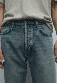 Faded blue denim jeans with classic five-pocket design, slightly distressed texture, and yellow stitching at the seams. Paired with a gray T-shirt.