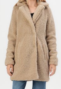 Béžový kabát z ovčí vlny s výstřihem do klopy, dlouhými rukávy a bočními kapsami. Materiál má měkký a strukturovaný vzhled.