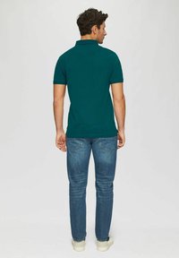 Dunkelgrünes Poloshirt für Herren mit kurzen Ärmeln, kombiniert mit mittelblauen Jeans und weißen Sneakers, von hinten betrachtet.