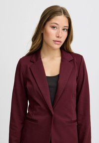 Blazer burdeos hecho de una tela suave. Presenta un cuello de solapa clásico, dos bolsillos frontales y un corte entallado.