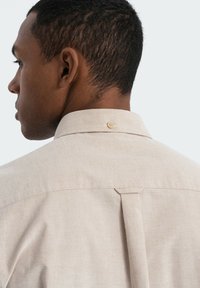 Beige linnen overhemd met een puntige kraag, een achterplooi en een enkele lichtgekleurde knoop bij de hals. Soepele textuur, casual ontwerp.
