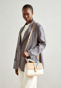 Sac à main beige clair et crème avec un rabat supérieur, quincaillerie dorée et une écharpe en soie bleue nouée sur la anse ; il présente un design structuré.