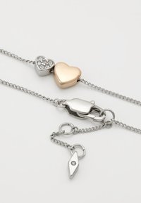 Collier en chaîne argentée avec un pendentif en forme de cœur en or rose et un plus petit cœur en argent incrusté de cristaux clairs, fixé par un fermoir homard en argent.