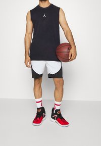 Maglietta nera senza maniche, pantaloni basketball bianchi e neri, scarpe da ginnastica rosse e nere, in mano una palla da basket, con calzini a righe rosse e bianche.