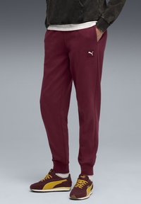 Bordeaux joggerbukser med et sort Puma-logo-patch. Blød bomuldsblanding, tætsiddende pasform, elastiske manchetter, parret med maroon og gule sneakers.