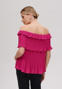 Rosa avskåret topp med plisserte stoffer, volangermede ermer og en flytende design. Båret med svarte bukser, som viser detaljene bak.