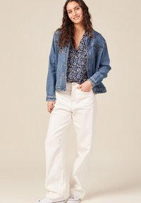 Giacca di denim sopra una blusa blu con motivo, abbinata a pantaloni bianchi a vita alta e larghi e sneaker bianchi. Accessori semplici inclusi.