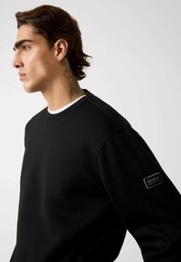 Czarna bluza wykonana z bawełnianej mieszanki. Posiada okrągły dekolt oraz dużą naszywkę z logo na rękawie, gładką fakturę i swobodny krój.