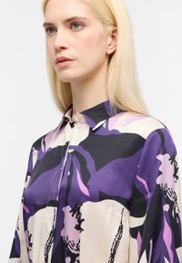 Blusa di seta con motivo floreale in viola, nero e crema; presenta un colletto a punta e chiusura a bottoni. Tessuto liscio e vestibilità comoda.