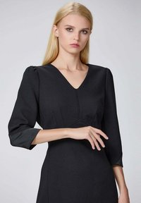 Robe noire avec un décolleté en V et des manches trois-quarts. Présente un corsage ajusté, des plis subtils et un tissu texturé.