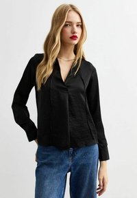 Blusa nera a maniche lunghe con scollatura a V e apertura frontale, realizzata in tessuto morbido. Abbinata a jeans in denim azzurri a vita alta.