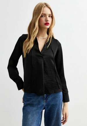 Zwarte blouse met lange mouwen en een V-hals, voorzien van een voorste knoopsluiting, gemaakt van een gladde stof. Gecombineerd met blauwe high-waisted spijkerbroek.