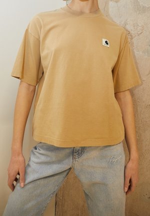 Person iført en løstsiddende beige t-shirt med et lille logo på brystet og lyseblå jeans med "Pegador" broderet på låret.