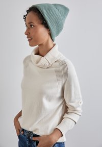 Maglione beige chiaro con colletto piegato e dettagli con bottoni, caratterizzato da accenti in maglia testurizzata sulle spalle. Abbinato a un berretto teal.