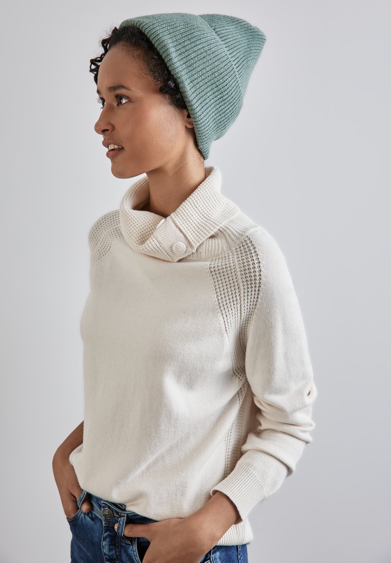 Maglione beige chiaro con colletto piegato e dettagli con bottoni, caratterizzato da accenti in maglia testurizzata sulle spalle. Abbinato a un berretto teal.