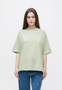 Lichtgroen T-shirt met korte mouwen, een hoge halslijn en een losse pasvorm. Draagt bij een blauwe jeans met slijtageplekken, met een gladde katoenen textuur.