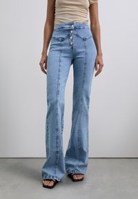 Vrouw in lichtblauwe high-waisted wijdpijpen jeans met knopen en een beige top, staand op een gladde grijze vloer tegen een witte muur.