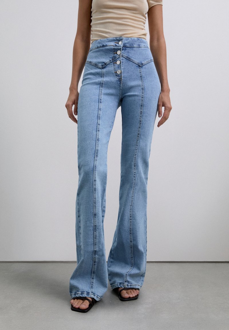 Vrouw in lichtblauwe high-waisted wijdpijpen jeans met knopen en een beige top, staand op een gladde grijze vloer tegen een witte muur.