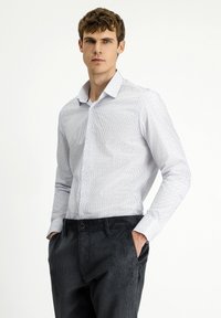 Herenbutton-up shirt in het wit met smalle blauwe strepen, gecombineerd met donkere corduroy broek. Het shirt heeft een gespreide kraag en lange mouwen.