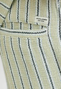 Tessuto intrecciato verde e beige con strisce verticali nere, bianche e blu e un'etichetta "JACK & JONES EST. 1990" cucita vicino a una tasca.