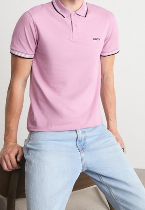 Mann trägt ein hellrosa Polohemd mit schwarz gestreiftem Kragen und Ärmeln, kombiniert mit hellblauen Jeans, sitzt auf einem Holzstuhl.