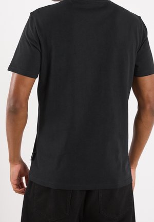 Mand set bagfra iført en simpel sort kortærmet T-shirt og sorte bukser mod en hvid baggrund.