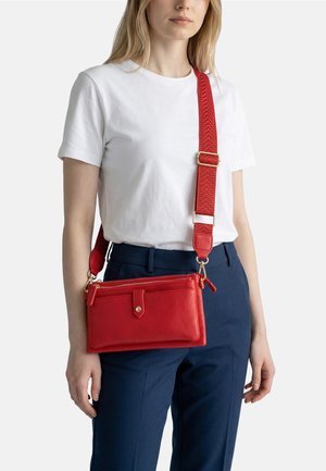 Donna che indossa una t-shirt bianca e pantaloni blu navy con una borsa a tracolla rossa in pelle, dotata di una tracolla larga regolabile e rifiniture in oro.
