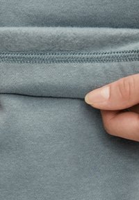 Lys blå hettegenserstoff med et teksturert fleeceinteriør. En persons hånd holder kantlinjen, som viser sydetaljene.