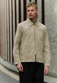 Chaqueta de cuero beige con cierre de cremallera, cuello y bolsillos laterales. Textura suave con un diseño aerodinámico, combinada con pantalones negros.