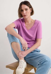 Tričko s výstřihem do V a krátkým rukávem v světle fialové barvě; spojené s modrými kalhotami uvolněného střihu a béžovými teniskami. Přírodní dřevěný stolička.