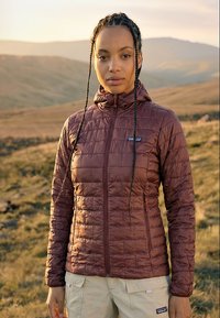 Kvinde med fletninger iført en vinrød quiltet jakke og beige cargobukser, der står udendørs med bakker i baggrunden.