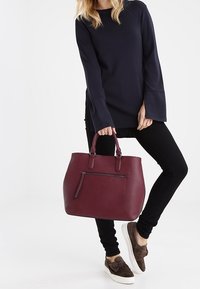 Bordeaux leren totebag met een afgeronde vorm, dubbele handvatten en een voorste ritszak. Gladde textuur en minimale hardware accenten.