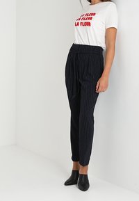 T-shirt blanc avec texte rouge, associé à un pantalon à rayures marines. Le pantalon a une taille haute et un détail de ceinture visible. Mules noires à talons.