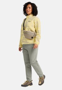 Veste en polaire jaune avec un motif subtil, sac bandoulière gris, pantalons verts et chaussures de randonnée noires et beiges. Le mannequin se tient contre un fond blanc.