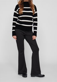 Un pull en maille à rayures noires et blanches avec un col haut, associé à un pantalon noir évasé et des chaussures en cuir verni. Tissu texturé.