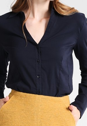 Mujer que lleva una camisa negra ajustada de manga larga abotonada, metida dentro de una falda texturizada color mostaza con las manos en los bolsillos.
