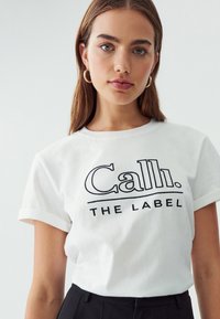 Valge puuvillane T-särk lühikeste rullitud varrukatega ja musta tekstiga "Call. THE LABEL" julge ja graafilise disainiga.