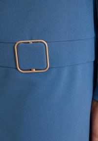 Blauer Stoffgürtel mit einer großen quadratischen beigen Schnalle um die Taille, ein Teil eines Kleidungsstücks neben einer Hand sichtbar.