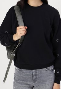 Personne portant un sweat-shirt noir avec de petits motifs en forme de cœur sur les manches, tenant la sangle d'un sac à bandoulière gris texturé, et portant un jean gris.