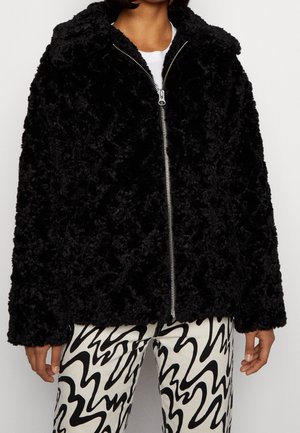 Person, die eine schwarze, flauschige Jacke mit Reißverschluss und schwarz-weiße abstrakt gemusterte Hose trägt, steht vor einem schlichten Hintergrund.