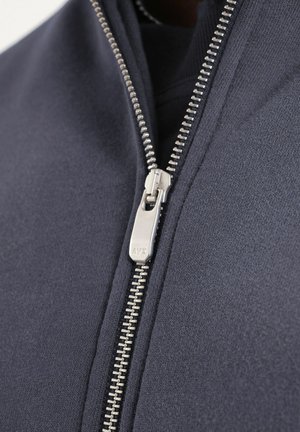 Plano detalle de una cremallera metálica plateada sobre una tela azul marino oscura, parcialmente cerrada con los dientes y la lengüeta del tirador visibles.