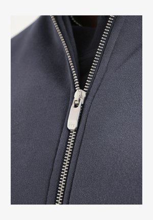 Plano detalle de una cremallera metálica plateada sobre una tela azul marino oscura, parcialmente cerrada con los dientes y la lengüeta del tirador visibles.