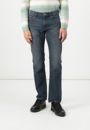 Personne portant un jean droit bleu foncé, des chaussures noires à lacets et un haut à manches longues rayé vert et blanc, debout devant un fond uni.
