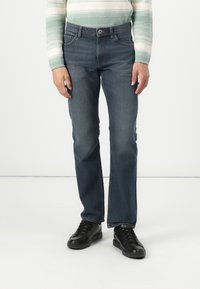 Jeans in denim blu scuro con un design a gamba dritta, dotati di cinque tasche e chiusura a bottone. Indossati con scarpe nere stringate.